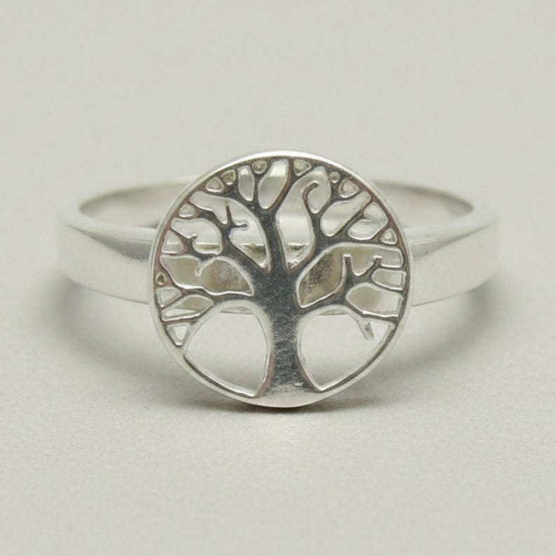 May include: A polished silver ring featuring a circular centerpiece with a cut-out design of a tree with spreading branches and a solid trunk. The band is smooth and tapers slightly toward the back, showcasing a metallic finish throughout.