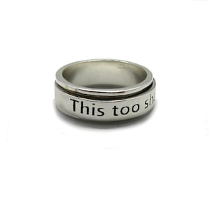 May include: A silver ring with the inscription "This too shall pass" in black lettering. The ring has a rotating outer band and a polished finish. The ring is set against a white background.