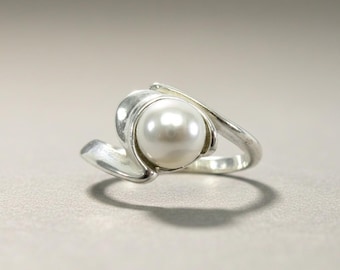 May include: A polished silver ring featuring a single, round, white pearl set in a bypass-style band. The metal band curves gracefully around the pearl, creating a smooth, continuous design that highlights the luster of the white gemstone.