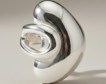 May include: A polished silver ring featuring a smooth, rounded, and asymmetrical organic shape. The design includes a prominent, curved outer surface that wraps around a central, hollowed-out opening, creating a continuous, reflective metallic form.