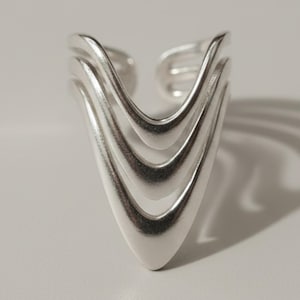 May include: A silver-toned open ring featuring a triple-layered V-shaped design. The band consists of three parallel, smooth, metallic arcs that converge into a single pointed tip, creating a geometric structure with an adjustable open back.