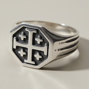 Peut inclure: Une bague chevalière en argent avec un plateau octogonal orné d'une croix de Jérusalem en relief. Le fond de la croix est oxydé en gris foncé, créant un contraste avec l'argent poli. L'anneau présente trois rainures horizontales.