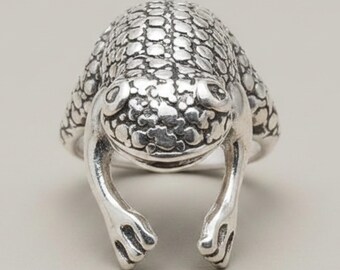 May include: A silver ring shaped like a frog, featuring a textured, bumpy surface across the head and back. The frog's front legs extend downward, forming the band of the ring, with clearly defined toes and eyes visible on the top of the head.