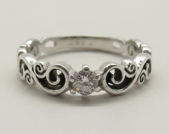 May include: A silver ring featuring a central round clear gemstone set in a prong setting. The band is crafted with a continuous series of raised scrollwork patterns that extend around the entire circumference of the metal ring.