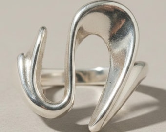 May include: A polished silver ring featuring an open, fluid, and abstract design with smooth, curved lines that create a continuous, wave-like shape. The metal has a reflective surface with soft highlights, forming an irregular, sculptural silhouette.