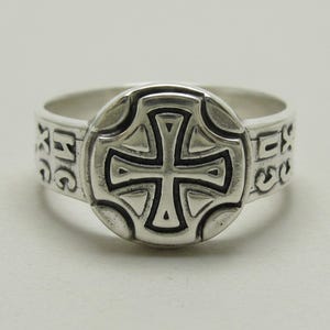 Könnte beinhalten: Ein silberner Ring mit einer kreisförmigen Vorderseite und einem erhabenen Kreuzdesign. Die Ringschiene ist auf beiden Seiten des zentralen Kreuzmotivs mit den Buchstaben IC XC graviert. Das Metall hat ein poliertes Finish mit dunklen Vertiefungen.