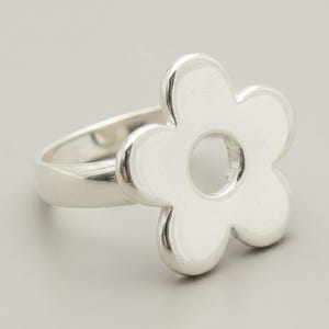 Könnte beinhalten: Ein Ring aus poliertem Silber mit einem markanten, fünfblättrigen Blumendesign. Das Blumenmotiv ist mittig auf der Ringschiene platziert und weist in der Mitte eine kreisförmige Aussparung auf, was eine glatte, reflektierende Oberfläche erzeugt.