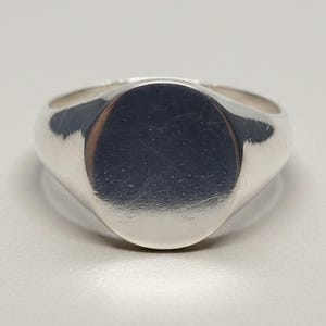 May include: A polished silver signet ring featuring a smooth, circular flat face and a solid, tapered band. The metal surface has a reflective, metallic finish with subtle light reflections across the rounded top and the band of the ring.