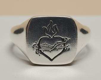 May include: A polished silver signet ring featuring a square face with an engraved design of a heart encircled by a crown of thorns and topped with a flame. The metal surface is smooth and reflective, highlighting the detailed dark etched lines.