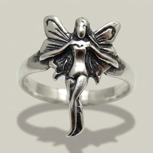 May include: A silver-toned metal ring featuring a central ornament shaped like a winged figure with outstretched arms and detailed wing patterns. The metal has a polished finish with darkened recessed areas to highlight the figure's form.