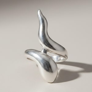 May include: A silver-toned ring featuring a fluid, organic shape with two elongated, curved forms that overlap and connect in the center. The metal surface has a smooth, polished finish that reflects light, creating highlights along the curves.