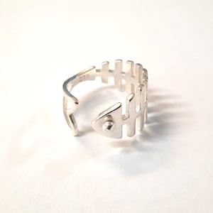 Puede incluir: Un anillo abierto de color plateado con un diseño de esqueleto de pescado. La banda tiene forma de una serie de costillas verticales que se extienden desde una columna central, con una pieza de cabeza triangular en un extremo. El interior tiene el sello 925.