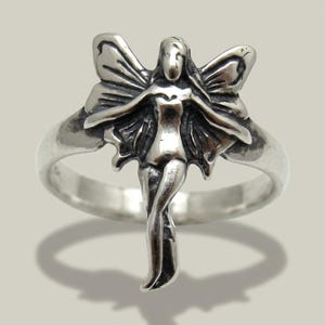 May include: A silver-toned ring featuring a central ornament shaped like a winged figure with outstretched arms and legs positioned together. The metal surface has a polished finish with darkened recessed areas to highlight the figure's details.