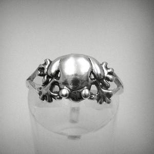 Puede incluir: Un anillo de tono plateado que presenta un elemento central liso y redondeado, flanqueado por estructuras intrincadas y ramificadas similares a hojas o coral en ambos lados. El metal tiene un acabado pulido y reflectante.