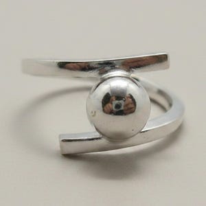 Könnte beinhalten: Ein polierter silberfarbener Ring mit offenem Banddesign. Der Ring besteht aus zwei parallelen horizontalen Stäben, die durch ein einzelnes, glattes, kugelförmiges Element in der Mitte verbunden sind und eine reflektierende Oberfläche bilden.