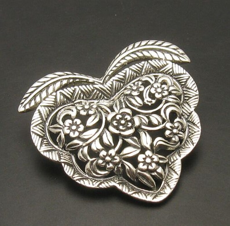 May include: A silver heart-shaped pendant featuring an intricate floral design. The pendant is crafted from delicate filigree work and showcases a beautiful floral pattern.