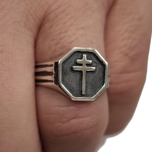 Peut inclure: Bague argentée avec une face octogonale ornée d'une croix. La bague a un anneau texturé avec des lignes horizontales. Le symbole de la croix est centré sur un fond gris foncé.