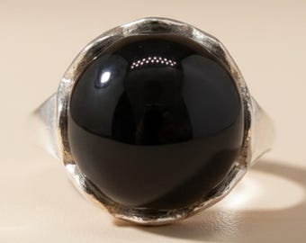 Op de afbeelding: Een zilveren ring met een grote, gladde, ronde zwarte steen in een zetting. De metalen band heeft een getextureerde, onregelmatige rand rondom de gepolijste zwarte cabochon, die het licht op het oppervlak reflecteert voor een contrastrijk effect.