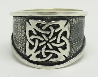 May include: A wide silver ring featuring a prominent, raised, polished silver knotwork design centered on a dark, textured, oxidized silver band. The band has smooth, polished silver edges at the top and bottom, creating a distinct contrast.