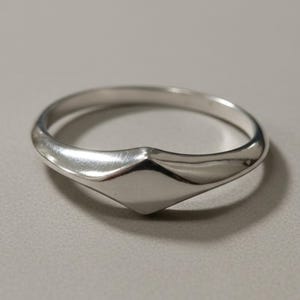 Op de afbeelding: Een gepolijste zilverkleurige ring met een gladde, doorlopende band die taps toeloopt naar een centraal, verhoogd ruitvormig punt. Het metalen oppervlak is reflecterend en uniform, met een strak, geometrisch ontwerp zonder extra stenen of graveringen.