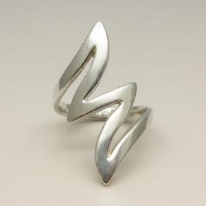Könnte beinhalten: Ein silberfarbener Ring mit einem einzigartigen Zickzack-Design. Der Ring hat eine Reihe von gebogenen, spitzen Formen, die einen kühnen, modernen Look erzeugen. Der Ring besteht aus einem glänzenden, metallischen Material.