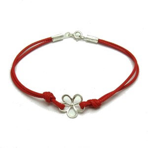 Könnte beinhalten: Ein rotes Kordelarmband mit einem silberfarbenen Metallblumenanhänger in der Mitte. Die Kordel ist auf beiden Seiten der Blume mit Knoten befestigt und verfügt über einen silberfarbenen Karabinerverschluss am oberen Teil des Armbands.
