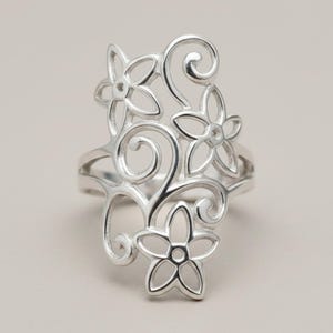 May include: A silver-toned ring featuring an intricate openwork design of three five-petaled flowers connected by swirling, curved metal lines. The metal has a polished, reflective finish, and the structure creates a tall, elongated shape.