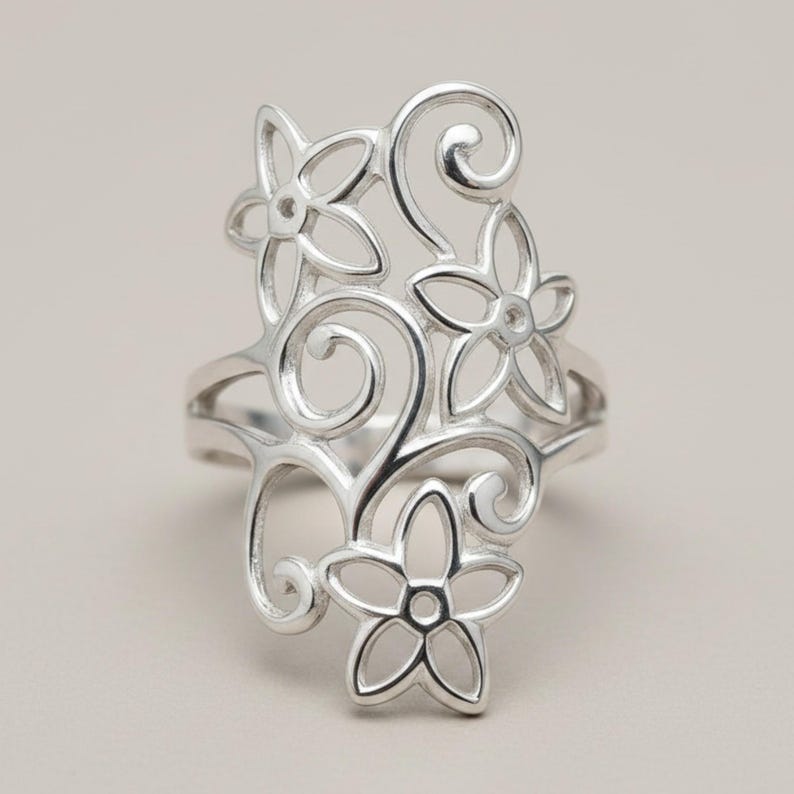 May include: A silver-toned ring featuring an intricate openwork design of three five-petaled flowers connected by swirling, curved metal lines. The metal has a polished, reflective finish, and the structure creates a tall, elongated shape.