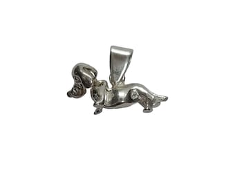 May include: A polished silver-toned metal pendant shaped like a dachshund dog in a standing position. The pendant features a smooth, reflective surface and includes a vertical bail at the top for attachment to a necklace chain or cord.