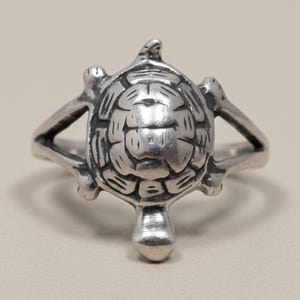 Op de afbeelding: Een zilverkleurige ring met een gedetailleerd schildpadmotief als middelpunt. Het schild van de schildpad heeft een gesegmenteerd, gestructureerd patroon en de ringband splitst zich in twee armen die aan de zijkanten van de schildpad vastzitten.
