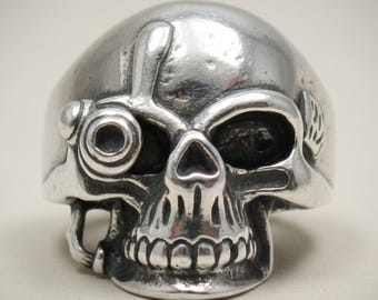 Può includere: Un anello in metallo color argento a forma di teschio umano. Il teschio presenta un prominente dettaglio circolare simile a un monocolo sopra l'orbita destra, una cavità nasale definita e denti visibili. La superficie ha una finitura lucida.