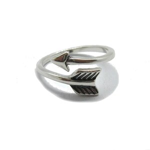 May include: A silver-toned metal ring shaped like an arrow that wraps around the finger. The ring features a pointed arrowhead on one end and a fletched tail end on the other, with oxidized detailing in the grooves to highlight the arrow design.