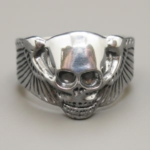 Peut inclure: Une bague en métal argenté poli présentant un motif central de crâne avec des orbites marquées et une mâchoire détaillée. Le crâne est flanqué d'ailes texturées et stylisées qui s'étendent le long des côtés de l'anneau, formant un bijou cohérent.