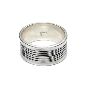 Può includere: Un anello d'argento con una fascia larga e un design testurizzato. La fascia è composta da più linee sottili che corrono attorno all'anello. L'anello è contrassegnato dal numero "925" all'interno.