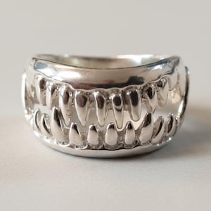 Könnte beinhalten: Ein Ring aus poliertem Silber mit einem durchgehenden Banddesign, das wie zwei Reihen spitzer, ineinandergreifender Zähne geformt ist. Die metallische Oberfläche ist glatt und reflektierend und bildet eine offene Struktur mit spitzen Fangzähnen.