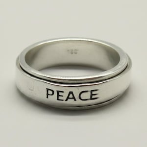 May include: A silver-colored metal spinner ring featuring a smooth, polished finish. The word PEACE is clearly engraved in black capital letters on the outer band. The interior of the ring band shows a small, faint hallmark stamp reading 925.