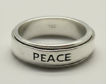 Puede incluir: Un anillo giratorio de metal color plata con un acabado liso y pulido. La palabra PEACE está grabada en letras mayúsculas negras en la banda exterior. El interior del anillo muestra un pequeño y tenue sello con el número 925.