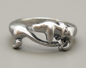 Op de afbeelding: Een zilverkleurige ring met een wikkelontwerp in de vorm van een teckel. De kop en voorpoten van de hond bevinden zich aan het ene uiteinde, terwijl het lange lichaam en de staart rondbuigen om de ring te vormen en de kop ontmoeten.
