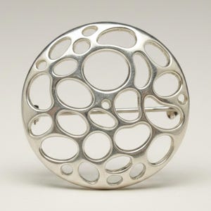 May include: A circular silver brooch featuring an openwork design composed of various sized, interconnected organic oval and circular cutouts. The metallic surface has a smooth, polished finish, and a horizontal pin mechanism is visible on the back.