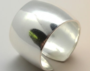 Puede incluir: Un anillo ancho de color plateado pulido con una superficie lisa y reflectante. La banda presenta un diseño abierto, creando un espacio entre los dos extremos. El metal tiene un brillo metálico que capta la luz, resaltando su forma curva.
