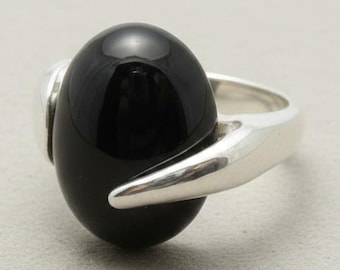 Puede incluir: Un anillo de plata pulida con una gran piedra negra lisa de forma ovalada en el centro. La banda de metal tiene un acabado liso y reflectante que se curva alrededor de la base de la gema oscura y brillante, creando un diseño sencillo.