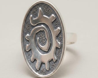 May include: An oval silver ring featuring a raised, central spiral design with radiating protrusions. The background of the oval face has a dark, textured, oxidized finish that contrasts with the polished, smooth surface of the silver spiral.