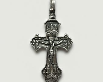 May include: A silver-toned metal crucifix pendant featuring an intricate relief design. The center depicts a figure on a cross, surrounded by smaller figures in the cross arms, a top panel with three figures, and a base panel with architectural details.