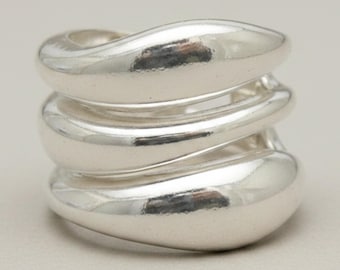 May include: A polished silver ring featuring a multi-layered design with three distinct, smooth, and rounded bands that wrap around the finger. The metallic surface is highly reflective, creating a continuous, fluid, and interconnected appearance.