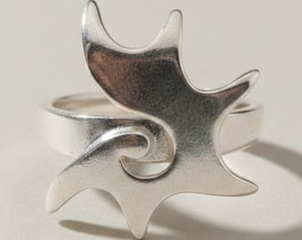 Könnte beinhalten: Ein silberner Ring mit einem markanten, flachen, stern- oder wellenförmigen Mittelstück. Das Metall hat eine glatte, polierte Oberfläche mit reflektierendem Glanz. Die Ringschiene ist kreisförmig und mit der Basis des Zierelements verbunden.