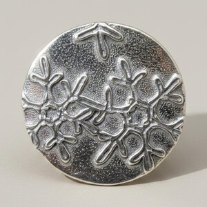 Puede incluir: Un pomo circular de metal plateado con un patrón de copos de nieve en relieve sobre su superficie. El diseño incluye múltiples motivos de copos de nieve grabados con ramas intrincadas sobre un fondo oscuro de textura punteada.