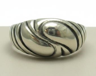 Op de afbeelding: Een gepolijste zilveren ring met een brede, bolvormige band met een reeks diepe, diagonale groeven die een wervelend, organisch patroon over het oppervlak creëren. Het metaal heeft een glanzende afwerking met donkere oxidatie in de lijnen.