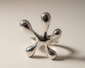 May include: A polished silver ring featuring a central organic, fluid shape with five rounded, bulbous protrusions extending outwards. The metal surface is smooth and reflective, creating a distinct star-like silhouette with a continuous band.