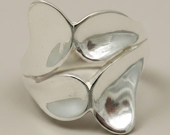 May include: A polished silver ring featuring a bypass design with two smooth, rounded, petal-like shapes that overlap slightly. The metal surface is highly reflective, creating soft highlights and shadows across the curved, organic forms of the band.