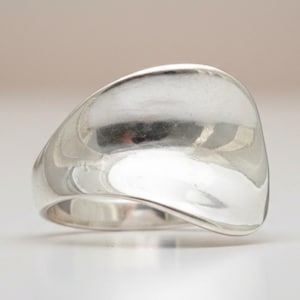 Puede incluir: Un anillo de plata pulida con una superficie ancha, lisa y curva que se estrecha en una banda delgada. El metal tiene un acabado brillante y reflectante con contornos orgánicos suaves que crean una forma fluida y continua.